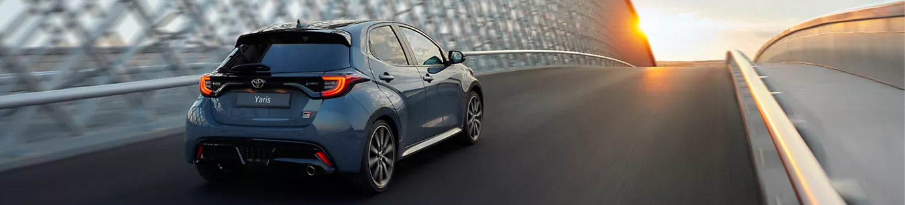 Toyota Yaris azul circulando por una carretera al atardecer, visto desde atrás